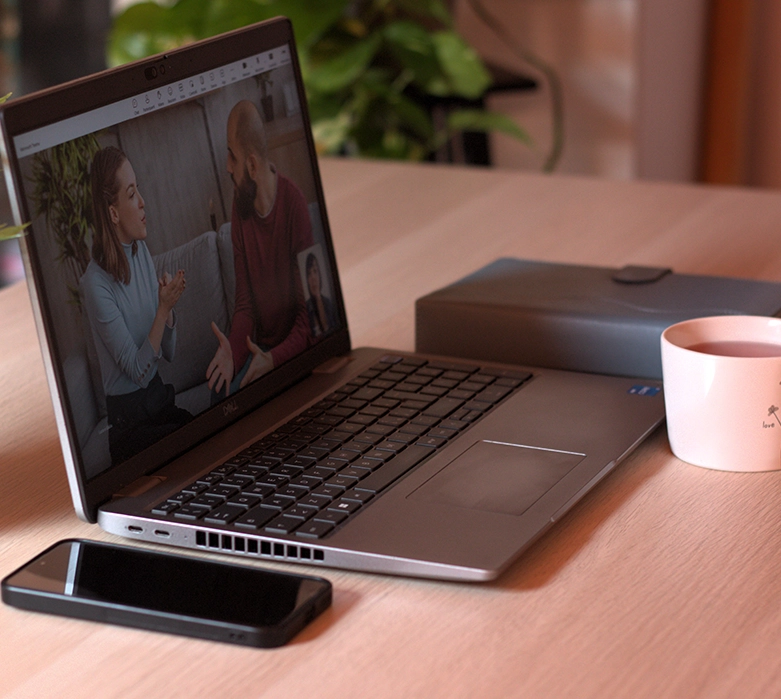 Laptop aperto su una scrivania durante una videochiamata di terapia e mediazione familiare online, con lo smartphone in primo piano e una tazza.