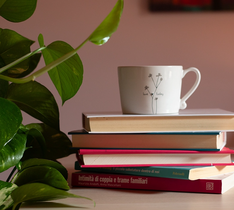Una pila di libri su relazioni e dinamiche familiari nello studio di Romina Schipano, sormontata da una tazza con scritto "love today" accanto a una pianta.