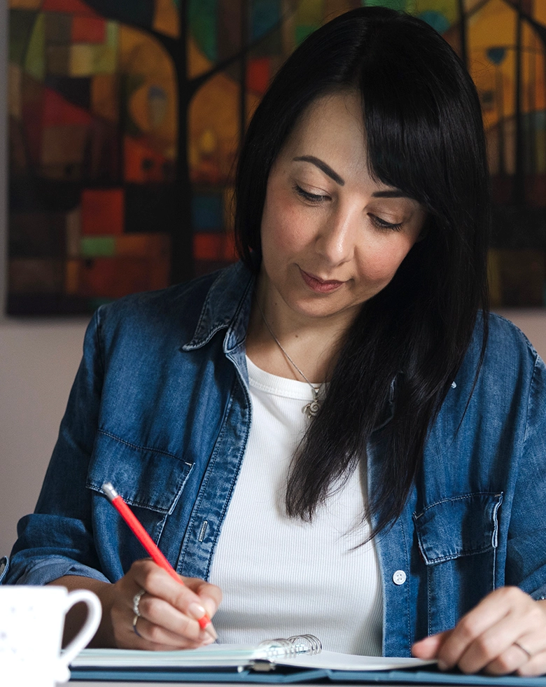 Romina Schipano, mediatrice familiare e titolare di Aiutarelazioni, seduta alla scrivania mentre scrive concentrata su un'agenda ad anelli con una matita rossa, indossa una camicia di jeans e sullo sfondo c'&egrave; un quadro colorato.